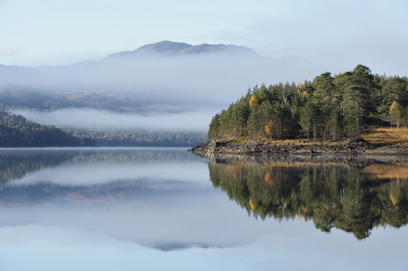 View over a loch with pine trees on the bank and mountains covered in mist in the background.