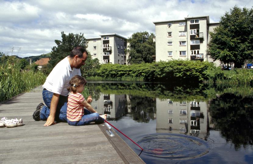 Adult and child pond dipping at the side of the canal with trees, plants and flats in background.