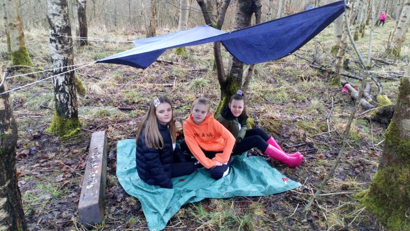 3 Primary 7 girls sitting on the forest floor under a tarpaulin tied to the trees