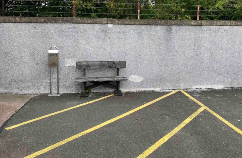 An area of asphalt and a wall at the end of the car park, with a small bench placed against the wall. 