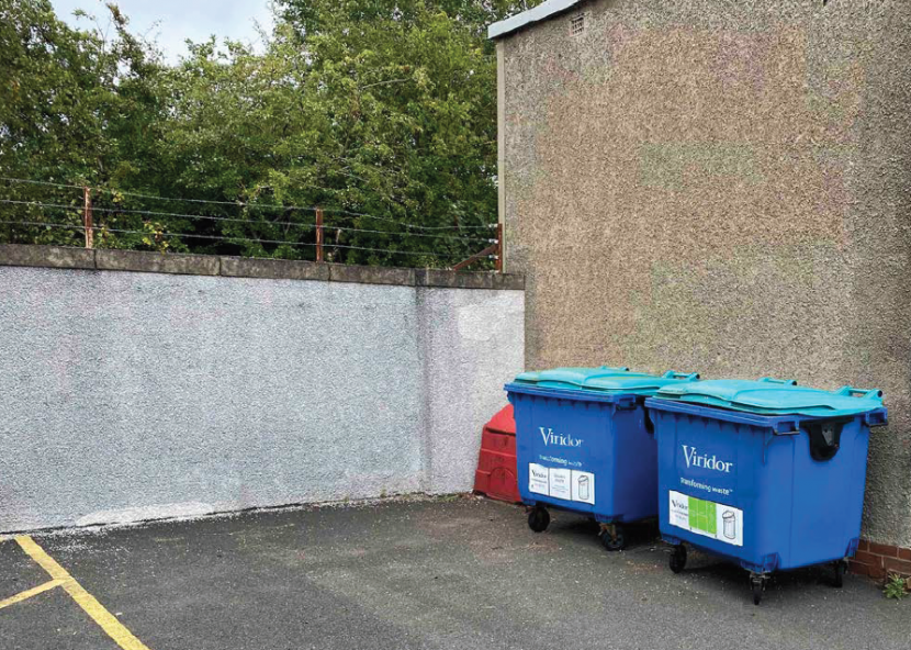 Two blue plastic wheelie bins placed against a wall at the eastern end of the asphalt covered car park.