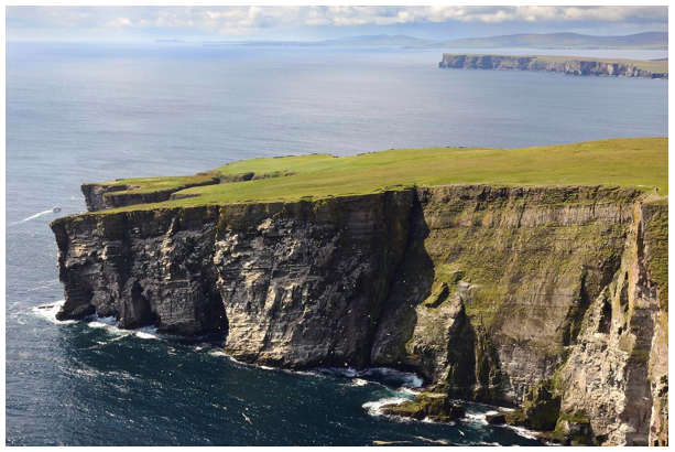 Plateau of  low green vegetation abruptly stops at a steep cliff plunging into the sea. 