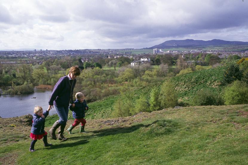 An adult and two young children walking near a loch, with a city and hills in the background.