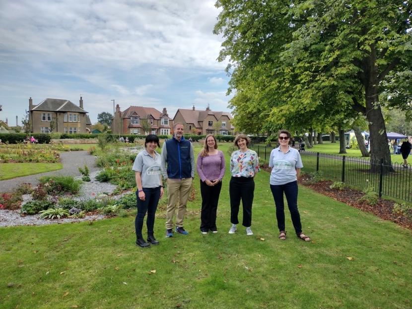Four people from various organisations that participated in the projects are standing in front of the raingarden's', surrounded by grass, trees and houses in the background.  