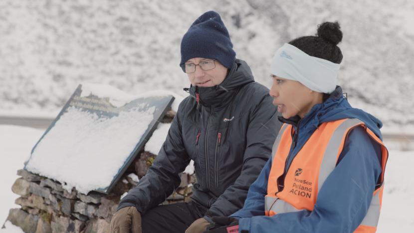 Two people sitign on a stone bench talking in the snow, wearing winter clothing and a high visibility vest.