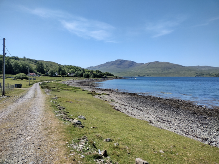 Shoreline on Mull, with pebble beach and mountains in the distance on a clear blue sky day.
