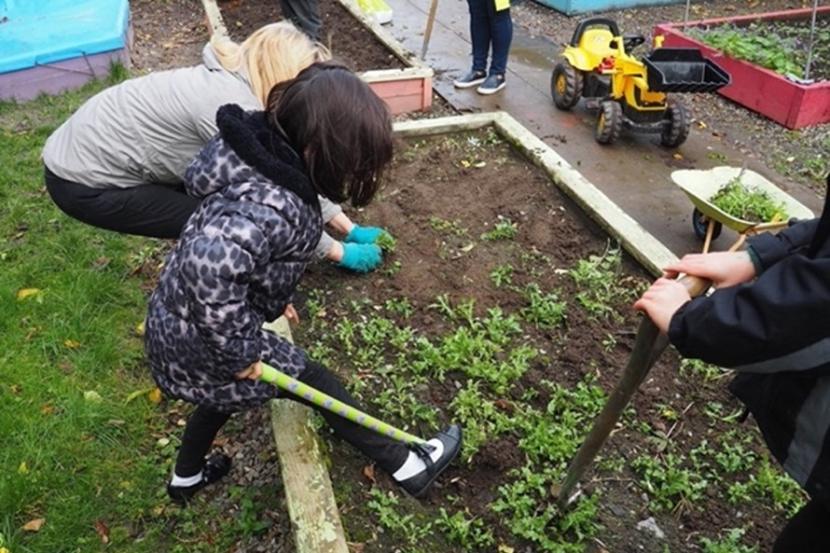 A child is digging in a raised bed alongside an adult. They are both wearing jackets, but the child is wearing white socks and open-topped patent leather shoes to dig with a small spade. ©&nbsp;Chris Mackie
