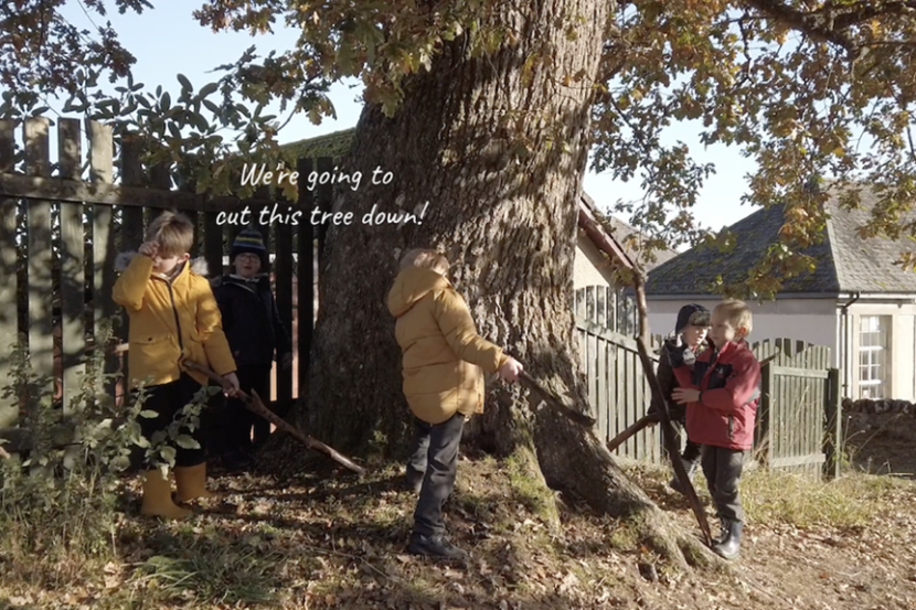 Five young children are gathered around mature oak tree on a bright, cold day. They are all holding sticks and taking turns to strike the tree trunk. A caption on the photo says, "We're going to cut this tree down!" © Chris Mackie