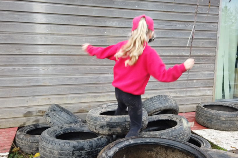 A child in school uniform is balancing their way over a pile of car tyres next to a timber-clad wall. Their hands are outstretched for balance, with one hand gripping a spindly tree branch. © Chris Mackie