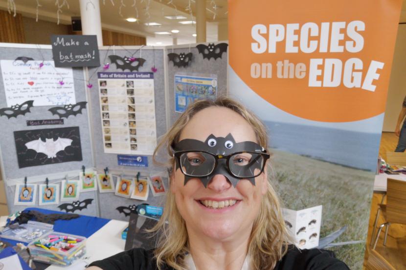 Person wearing a bat mask standing in front of a stand with arts and crafts items for making bat masks.