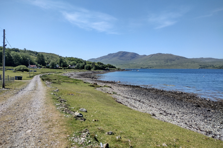 Shoreline on Mull, with pebble beach and mountains in the distance on a clear blue sky day.