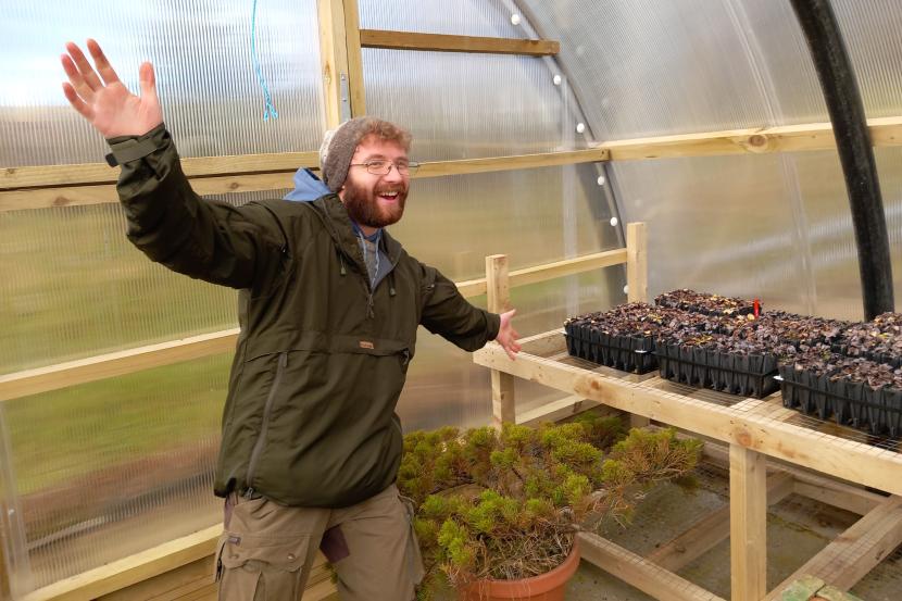 A person in outdoor clothing, in a greenhouse with trays of seedings on a bench behind them.