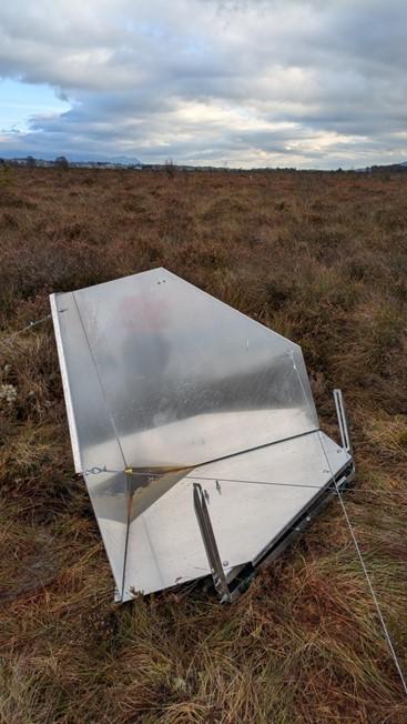 A large metallic reflector made of three connected sheets of aluminium slightly raised above the very low vegetation on the surface of bog.