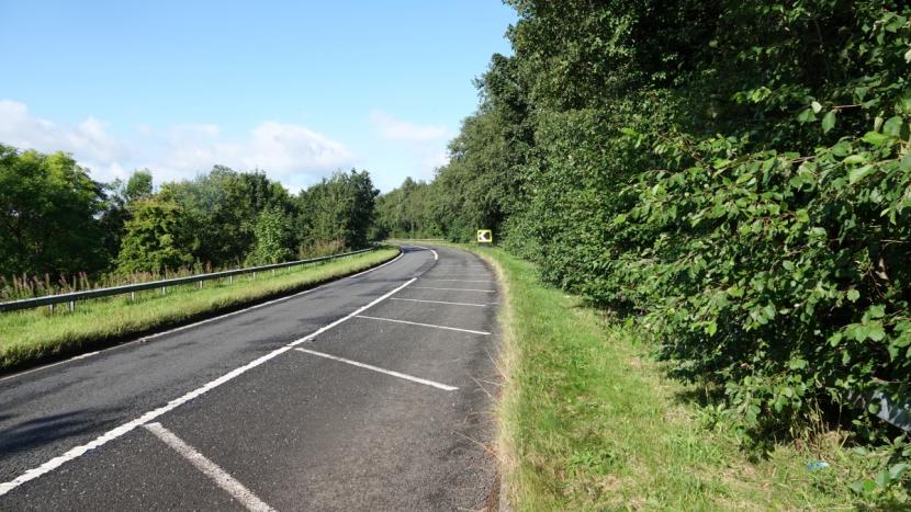 A single-lane off-slip road with narrow grassy verges bounded by woodland on the right and wider verge, safety barrier and liner woodland and scrub belt on the left. 
