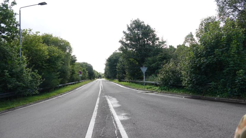 A two-lane access road to and from A82 with narrow green verges, safety barrier and tree-lined on both sides.