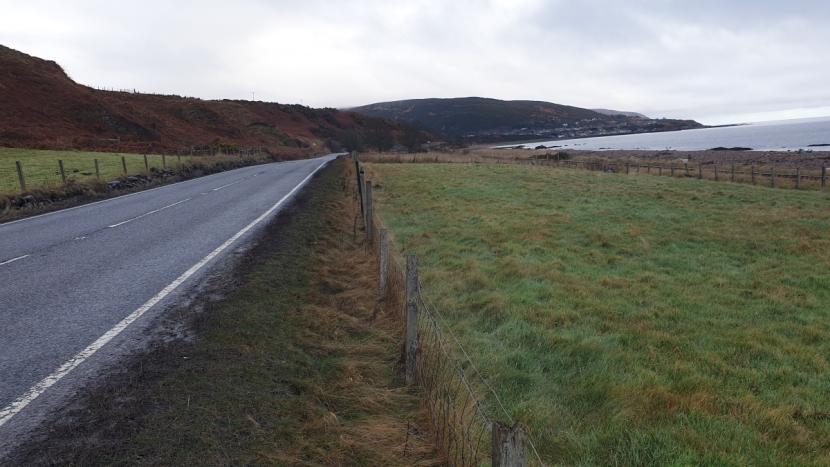 The A9 road northbound with stock fencing close to the road on both sides, and with pastures and hillsides on the left, and pastures, railway line and the coast to the right.