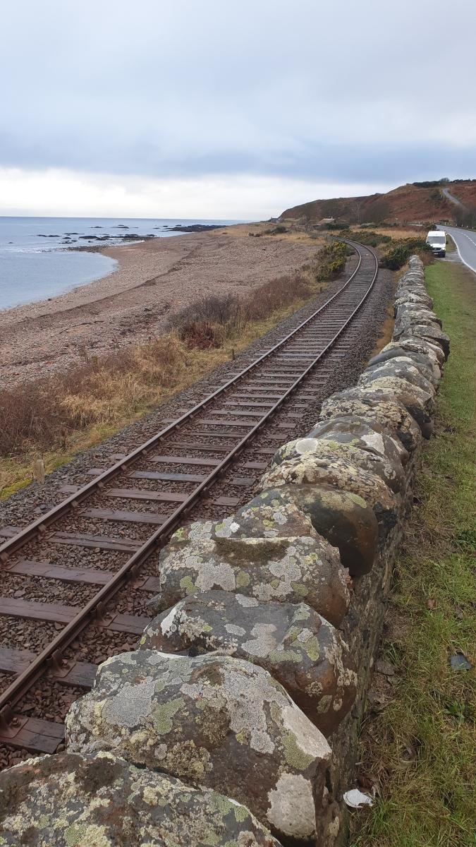 A railway line running within 10 m. of the A9 road in the foreground, with a low stone wall between them. 
