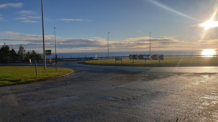 A roundabout with short grass within the centre, wide grassy verge on the left, and the sea in the background. 