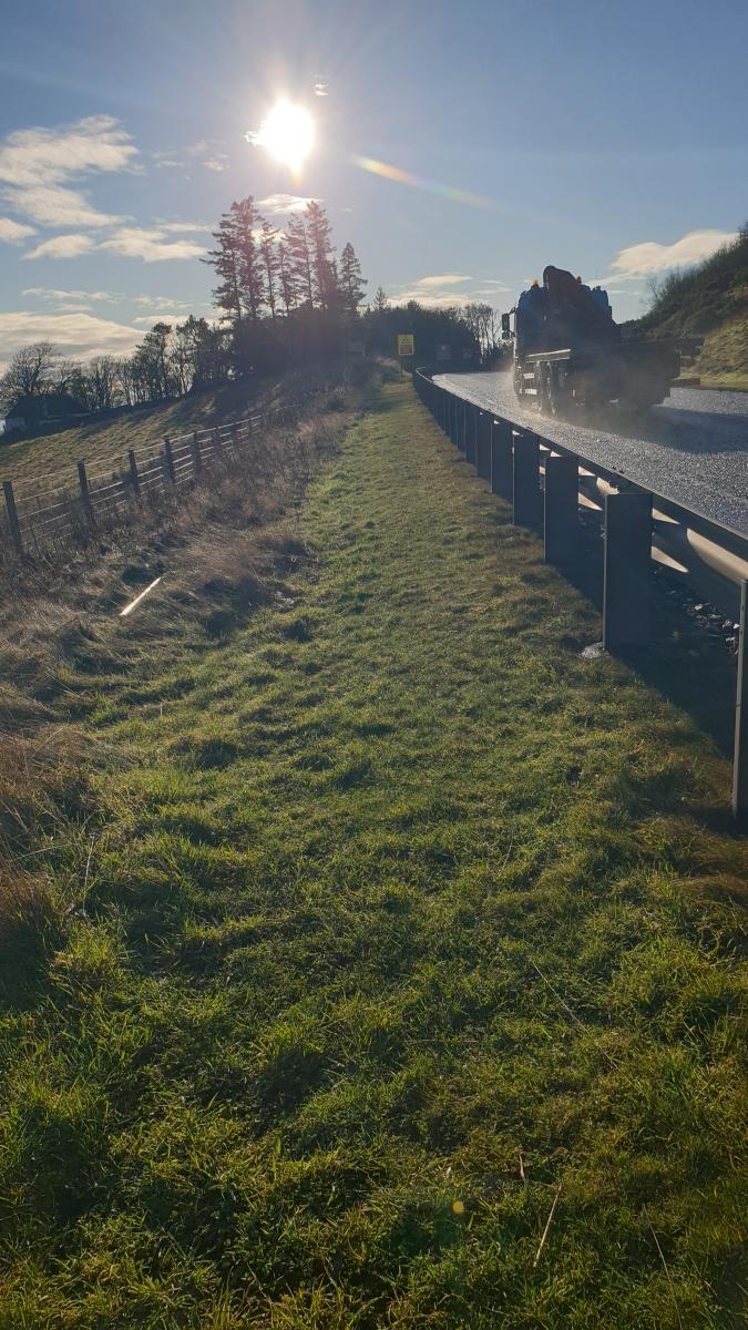 From left to right a line of stock fencing, a wide grass verge, a safety barrier and the A9 southbound. 