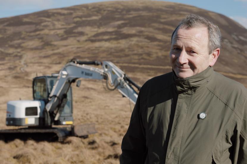 A man standing on a hillside with a digger behind.
