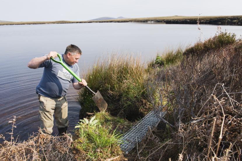 A man digging near a small cage on the banks of a loch. 