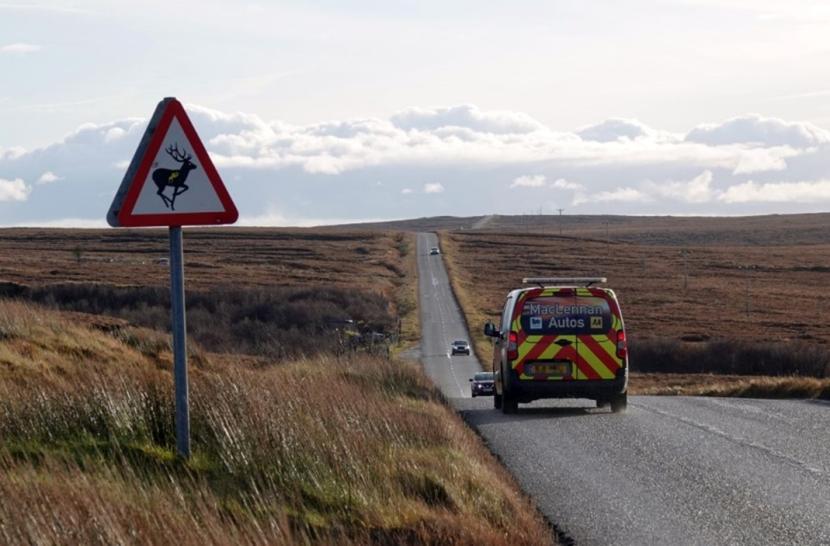 A typical view from the A857 leading through the undulating open landscape of Barvas Moor with intermittent blind summits and a wild animal warning sign. There are vehicles moving in both directions on the road. 