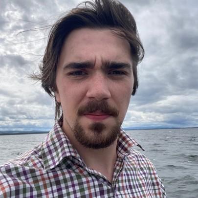 A selfie of a young man outside with a coastal landscape behind him.