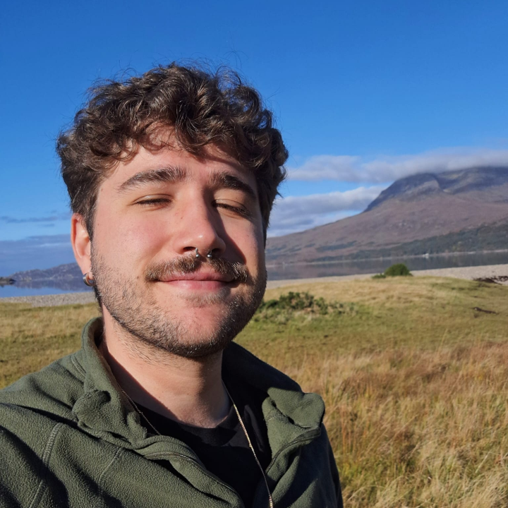 A selfie of a young man standing outside with hills and a blue sky in the background. 
