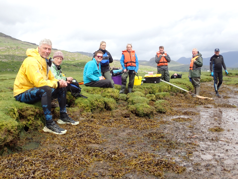 A group of people wearing outdoor gear and life jackets gathered along a grassy, rocky shoreline next to a body of water.