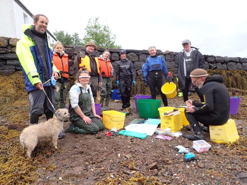A group of nine people wearing outdoor gear, and a dog on a rocky shore next to a stone wall. 