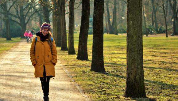Female walking in park smiling and wrapped up warm with jacket/scarf, with kids on bike in background