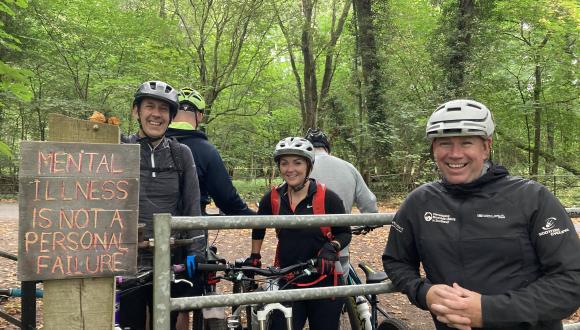 Three people outdoors, looking at camera, smiling, with bikes. Sign on fence posts reads 'mental illness is not a personal failure'. Image courtesy of Cycling Scotland
