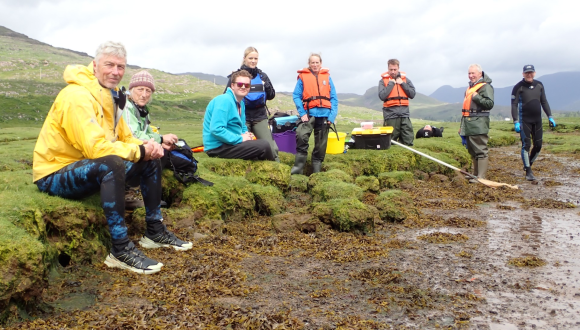 A group of people wearing outdoor gear and life jackets gathered along a grassy, rocky shoreline next to a body of water.