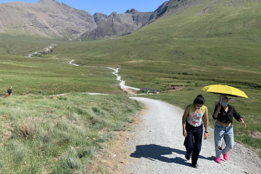 Two people walking on the Fairy Pools path; the Cuillin mountains in the background; one person with a yellow umbrella