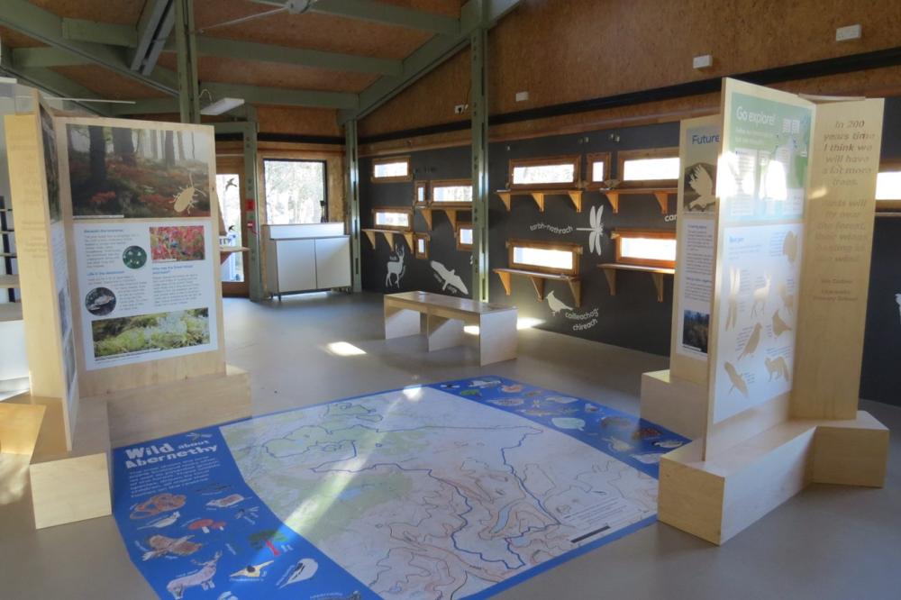 the interior of the Loch Garten Osprey centre, showing horizontal narrow windows for bird watching, a map on the floor, and interpretation panels left and right.