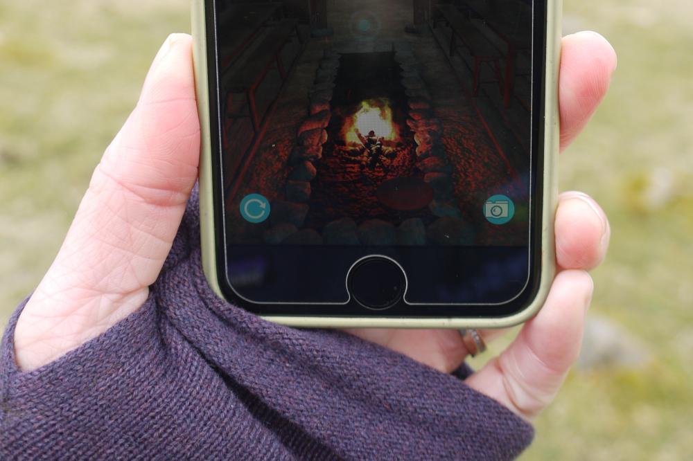 Uist Unearthed's app, on the Bornais site, showing a hand holding a phone, with the augmented reality image of a Viking Longhouse, and central fire.
